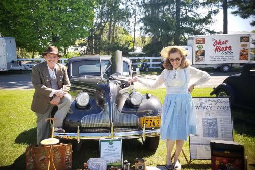 A man and woman in 1930s period dress pose with a black antique car.