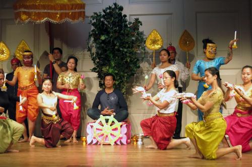 Dancers pose on stage; the women are in traditional skirts and blouses, but the men are mostly in jeans and shirts. A man sits cross-legged in the center on a small podium.