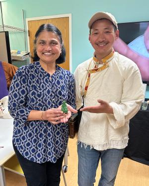 A woman holds up a small, green wax figure of the Buddha, while a man poses next to her, gesturing to the sculpture. They both smile.
