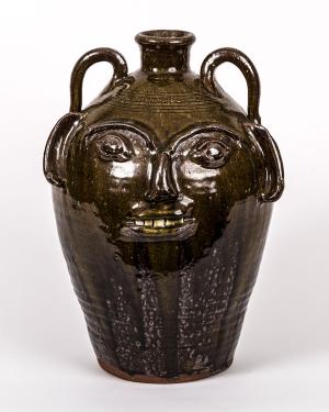 Bulbous dark jug with two curving handles near the mouth and a human-like face on its side, including protruding white teeth.