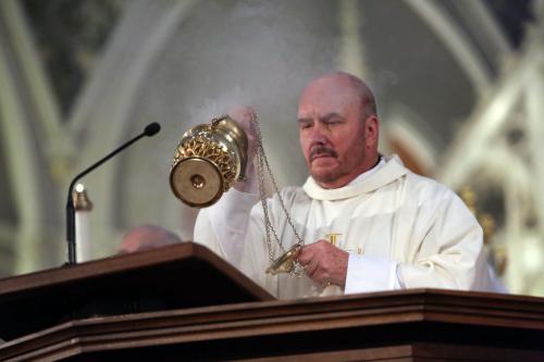 A Catholic priest dressed in a white robe waves a golden incense holder. A thin cloud of smoke is seen coming out of it.