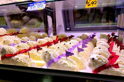 Display of various butter lambs in a refrigerated case. They have red or purple bows and small plastic flags in their backs. The 10 ounce size is priced at $9.99.