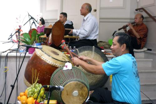 Musicians rehearse at the side of a stage. The man in front has two barrel drums and a gong, plus an offering of (possibly plastic) fruit.