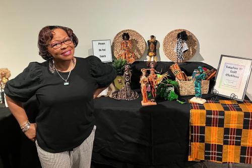 A woman in a black top and silver jewelry smiles, standing beside a display table of Barbie-like dolls wearing clothing made from African prints.
