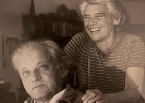 Black-and-white photo of a man, seated, and a woman standing behind him, hands on his shoulders, laughing.