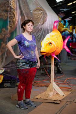 A woman with paint splattered all over her clothes poses next to a sculpture of a tropical fish, on a stand so it's as tall as her.