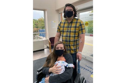 A woman in a wheelchair holding a new baby and man behind her pose at the entrance to a hospital.