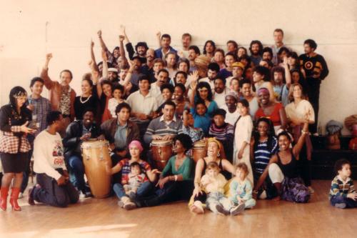 Dozens of men, women, and children pose in several rows against a white wall. Three men sit in front of wooden conga drums. Many people hold one fist in the air.