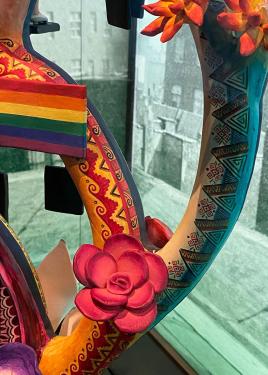 A close-up on the sculpture shows the rainbow pride flag, a pink flower, and intricate geometric patterns on the tree’s branches.