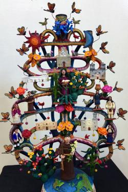 A multicolored clay sculpture in the abstract shape of a tree with curving branches and the Northern Hemisphere of Earth as the base. On the branches are figures of monarch butterflies, churches, people, cacti, and flowers.