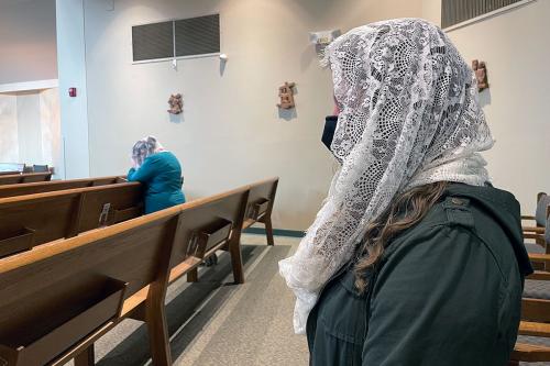 From the side, a woman stands at a church pew, her face obscured by a white lace veil over her head. Another woman in the background, bent over in prayer, wears the same type of veil.