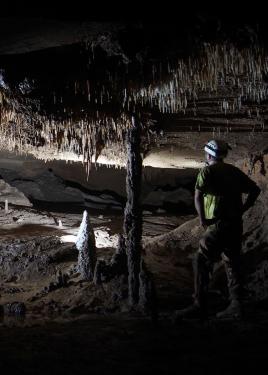 A person in hard hat and yellow safety vest looks out over an underground cavern with dramatic stalactites and stalagmites.