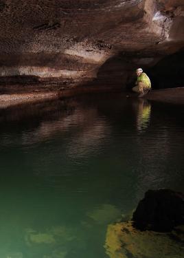 A person in hard hat and yellow safety vest crouches at the edge of an underground pool of water.