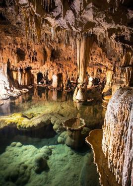 An illuminated pool of turquoise water in a cavern, with stalactites hanging down and tubular rock formations.