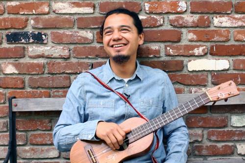 César poses with his jarana against a brick wall, smiling.