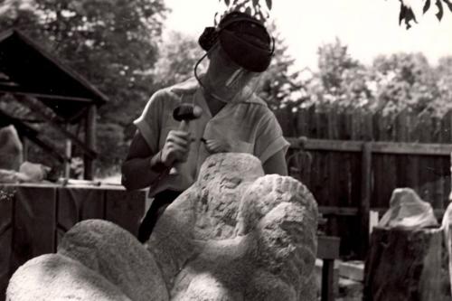 A woman stands in an outdoor studio holding a mallet and chisel, which she uses on the large stone sculpture in the foreground.