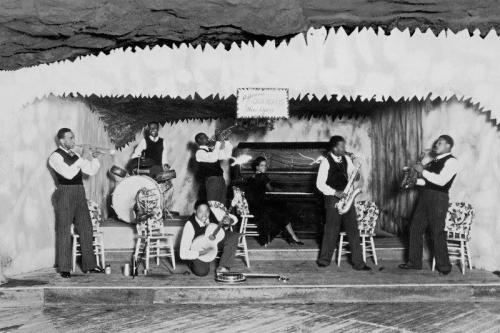 Seven musicians playing on brass instruments, acoustic guitar, drums, and piano, on a stage in a cave.
