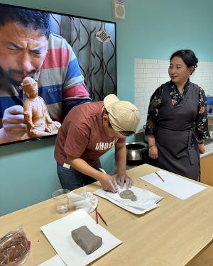 A man hunches over a table forming a piece of gray clay. Standing next to him is a woman, who looks over at a TV behind them, showing a photo of an older man forming a small sculpture of a person.