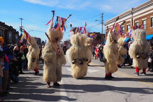 Figures in sheepskins and ribbon headdresses, with large metal bells attached at their waistlines, parade down a crowded street under blue skies.