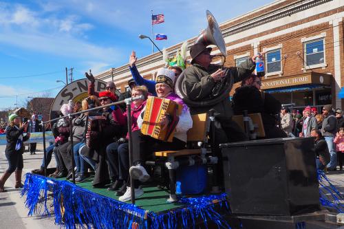 A parade float on a city street, holding people seated, waving to the crowd and playing accordions and a sousaphone.