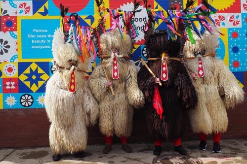 Four figures in sheepskin costumes—two off-white and one dark—pose infront of a colorful wall. They each  have ribbon headdresses, wooden clubs, and red ornaments representing tongues.
