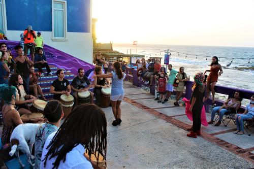 The community dance plaza at sundown alongside the ocean, with drummers along the sides and a dancer in front of them, and several kids waiting.