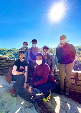 Six people wearing hiking gear and facemasks pose along a low stone wall, with a bright blue sky and forested landscape behind them.