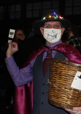 A person in purple dress shirt and tie, dark best, magenta cape, and hat with Christmas lights around the brim holds up a sticker that says NAUGHTY in one hand, and a wicker basket in the other.