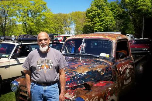 A man poses next to an old truck with layers of blue, white, and red paint scraped away, plus brown rusty spots. 