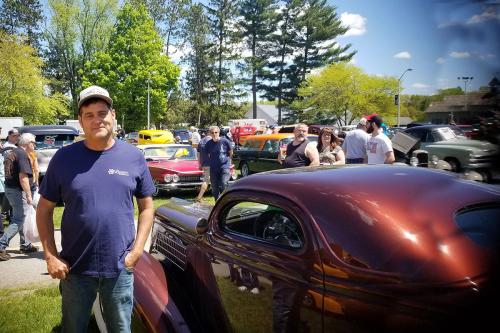 A man poses next to a short maroon car.