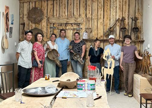 Eight people pose side by side. Two hold up wooden musical instruments, with strings strung over a carved bowl shape. Other instruments are displayed on the walls around them.