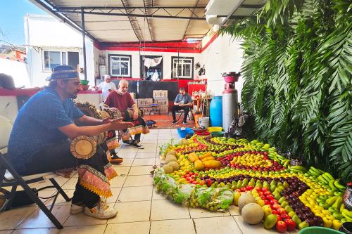 On a covered porch, two seated drummers face an altar of carefully arranged fruits and a backdrop of green palm leaves.