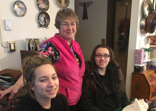 A grandmother in pink vest stands between two young women, seated, inside a home.