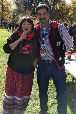A woman, wearing a black sweatshirt with a red graphic for Red Lake Nation and a long, red ribbon skirt, and holding a microphone up to her mouth, poses outdoors next to a man wearing a striped dress shirt, black embroidered vest, and jeans.