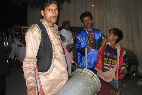 A nighttime wedding procession. A man in front carries a metal barrel-shaped drum over his shoulder and glares at the camera. Others in the background carry drums and brass instruments.