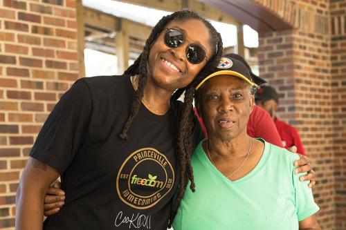 A young person with sunglasses, long locs, and a Princeville Homecoming T-shirt poses embracing an older woman wearing a Pittsburgh Steelers hat and a turquoise top.