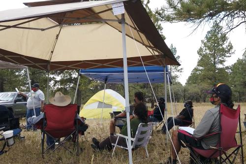 A mgroup of people meet outdoor under pop-up tents, wearing masks and physically distanced, in a field under trees.
