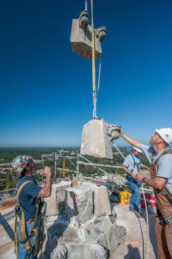 Two men in the foreground, wearing hardhats and safety harnesses, guide a crane in lifting stone pieces from the top of the cathedral. In the background, a third man, wooden scaffolding with metal railings, a view of Washington covered in green trees, and