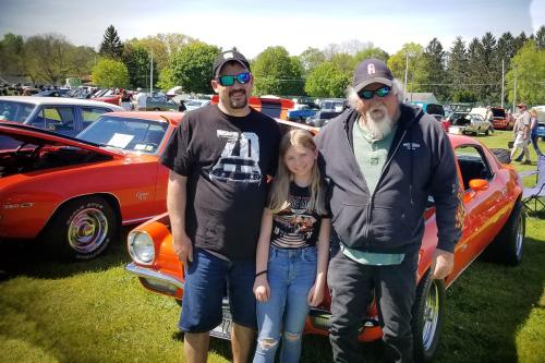 Two men and a young girl pose in front of a classic car.