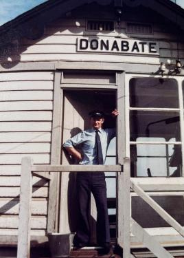 A man in a blue uniform poses in the doorway of a train station. Above him is a sign that reads Donabate. Old color photograph.