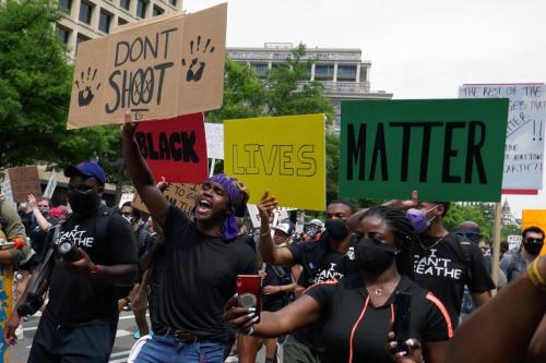 A group of people march through the street, some wearing masks. One is shouting, holding a sign that reads DONT SHOOT with two handprints and X eyes in the O’s of “SHOOT.” Other signs read BLACK (in red), LIVES (in yellow), MATTER (in green).