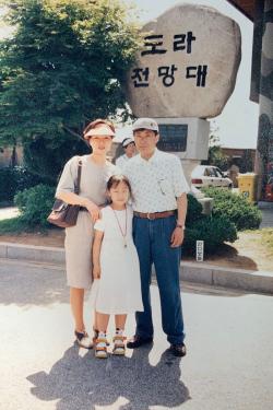 Two parents and young daughter pose, standing in front of an entrance sign, Korean characters carved into rock.