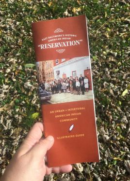 Closeup of a hand holding up a brochure, titled East Baltimore’s Historic American Indian “Reservation.” An Urban + Intertribal American Indian Community. Illustrated Guide. The photo shows a large group of adults and children posed on the sidewalk in fro