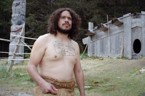 Film still of a man, shirtless with a tribal tattoo of a creature on his chest, looking beyond. Behind him are a wooden structure and a totem pole.