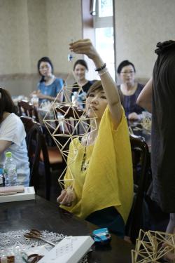 A woman in a workshop holds up a simple straw mobile. Other women behind her are working at long tables.