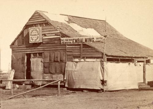 Ramshackle wooden building with a banner out front which reads, Embalming. Doctor Bunnell.