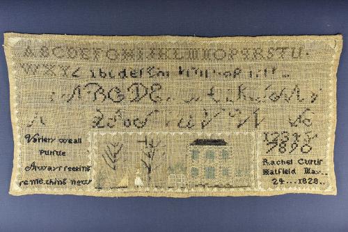 Old piece of cloth which someone used to practice embroidering the alphabet and numbers, a few phrases, and a design of a house with people and trees in the yard. Embroidered year reads 1828. Laid on a blue backdrop.