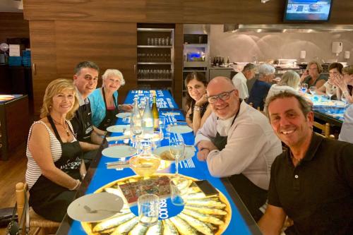 Six people smile for the camera, sitting at an illuminated table filled with plates and silverware, wine bottles and glasses.