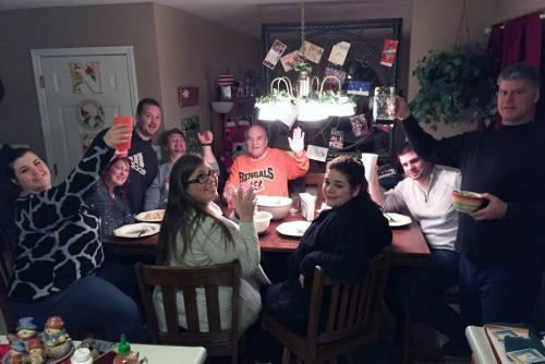 Around a square wooden tables, nine adults raise their glasses and wave toward the camera.