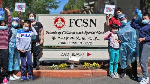 Kids and adults in face masks and face shields pose around a sign for FCSN: Friends of Children with Special Needs. 2300 Peralta Blvd. The sign also has the name in Chinese characters. 
